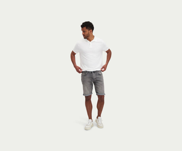 Een man met een wit poloshirt, The N01 Short Light Grey Used by Blue Rivet Jeans, en witte sneakers staat tegen een effen witte achtergrond. Hij kijkt naar voren met zijn handen op zijn heupen en kijkt lichtjes opzij.