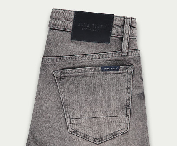Een paar lichtgrijze jeans netjes opgevouwen, met een achterzak met een klein label met de tekst Blue Rivet Jeans. Een zwarte leren patch siert de tailleband, wat een vleugje elegantie toevoegt aan deze veelzijdige denim essentials die bekend staan als The N01 Short Light Grey Used.