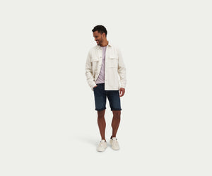 Man gekleed in een lichtbeige jas, een gestreept overhemd, The N01 Short Blue Black van Blue Rivet Jeans en witte sneakers, staat lachend tegen een effen achtergrond.