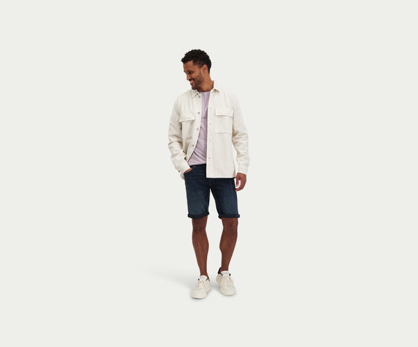 Man gekleed in een lichtbeige jas, een gestreept overhemd, The N01 Short Blue Black van Blue Rivet Jeans en witte sneakers, staat lachend tegen een effen achtergrond.