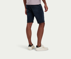 Een persoon die The N01 Short Blue Black van Blue Rivet Jeans draagt, gecombineerd met een wit T-shirt en witte sneakers, staat tegen een effen achtergrond. De afbeelding benadrukt de achterkant, toont de gevouwen zomen van de shorts en benadrukt de relaxte stijl van de outfit.