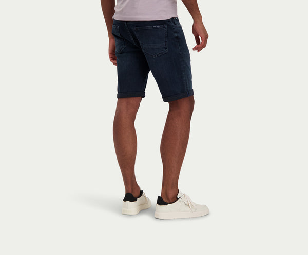 Een persoon die The N01 Short Blue Black van Blue Rivet Jeans draagt, gecombineerd met een wit T-shirt en witte sneakers, staat tegen een effen achtergrond. De afbeelding benadrukt de achterkant, toont de gevouwen zomen van de shorts en benadrukt de relaxte stijl van de outfit.