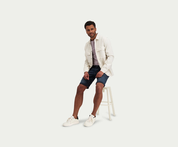 Een man zit op een witte kruk en draagt The N01 Short Blue Black van Blue Rivet Jeans. Hij combineert het met een lichtgekleurd overhemd met knopen over een t-shirt en witte sneakers. De effen witte achtergrond accentueert zijn relaxte maar stijlvolle look.