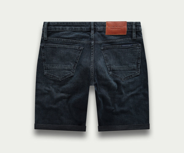 De N01 Short Blue Black van Blue Rivet Jeans is een zwarte denim short met opgerolde pijpen, twee achterzakken en een bruine leren patch op de tailleband tegen een lichte achtergrond.