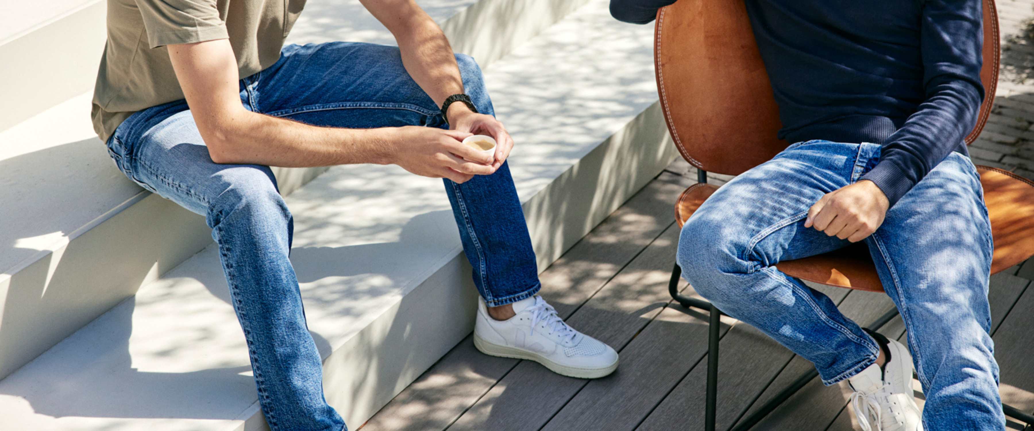 Twee mensen in spijkerbroek en sneakers zitten buiten – de een op een witte trap met een beker in de hand, de ander op een bruine stoel. Zonlicht en schaduwen van bomen zijn zichtbaar op de grond en de achtergrond. Gezichten zijn niet te zien.