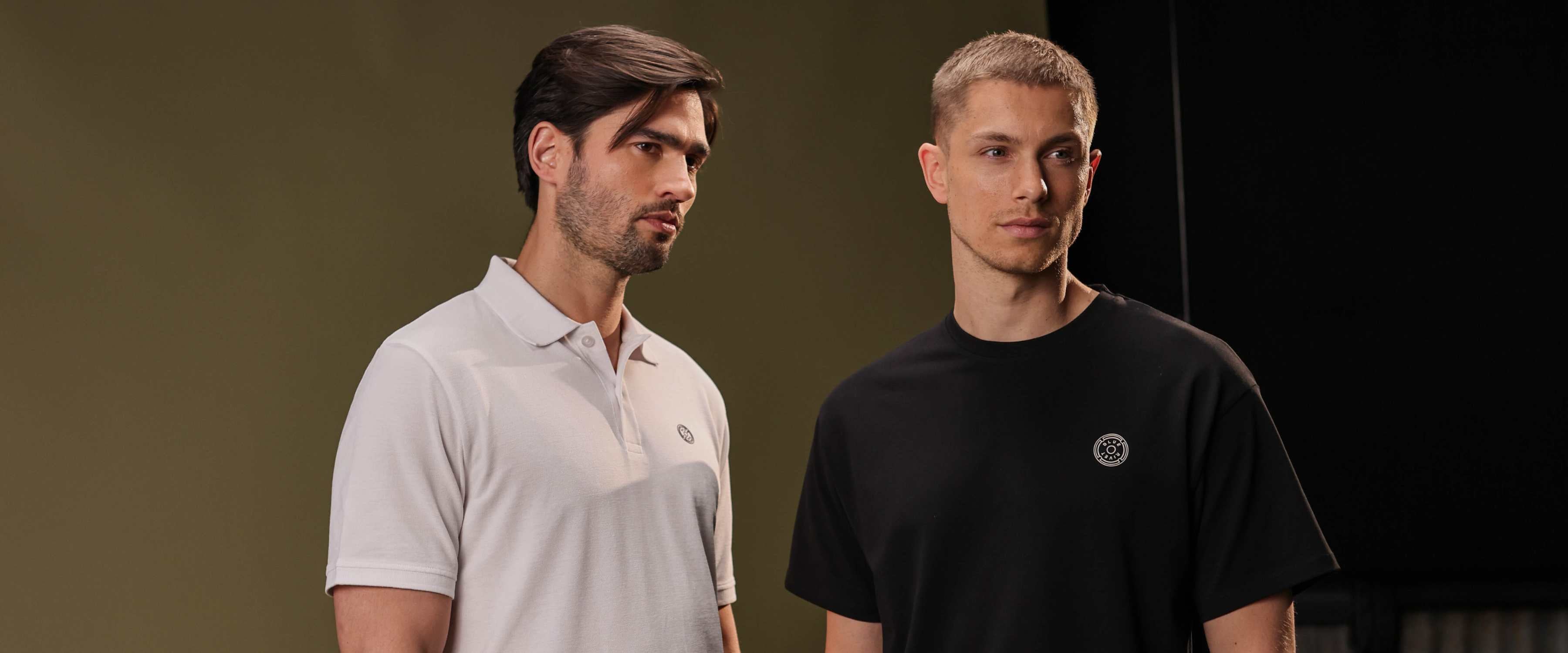 Een man met donker haar en een baard zit zijdelings op een houten loungestoel, draagt een zwarte polo, blauwe jeans en zwarte sneakers, tegen een neutrale achtergrond.