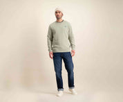 Een man gekleed in een lichtgrijze trui, The N01 Straight Dark Indigo Used jeans van Blue Rivet Jeans, witte sneakers en een beige muts staat tegen een effen beige achtergrond. Zijn houding is ontspannen met zijn handen langs zijn zijden terwijl hij lichtjes naar de zijkant kijkt.