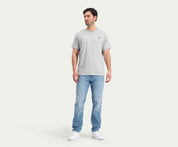 Een persoon staat tegen een effen, lichte achtergrond en draagt een grijs T-shirt, The N01 Straight Stone Bleached jeans van Blue Rivet Jeans en witte sneakers. Ze kijken iets opzij, met hun handen ontspannen langs hun lichaam.