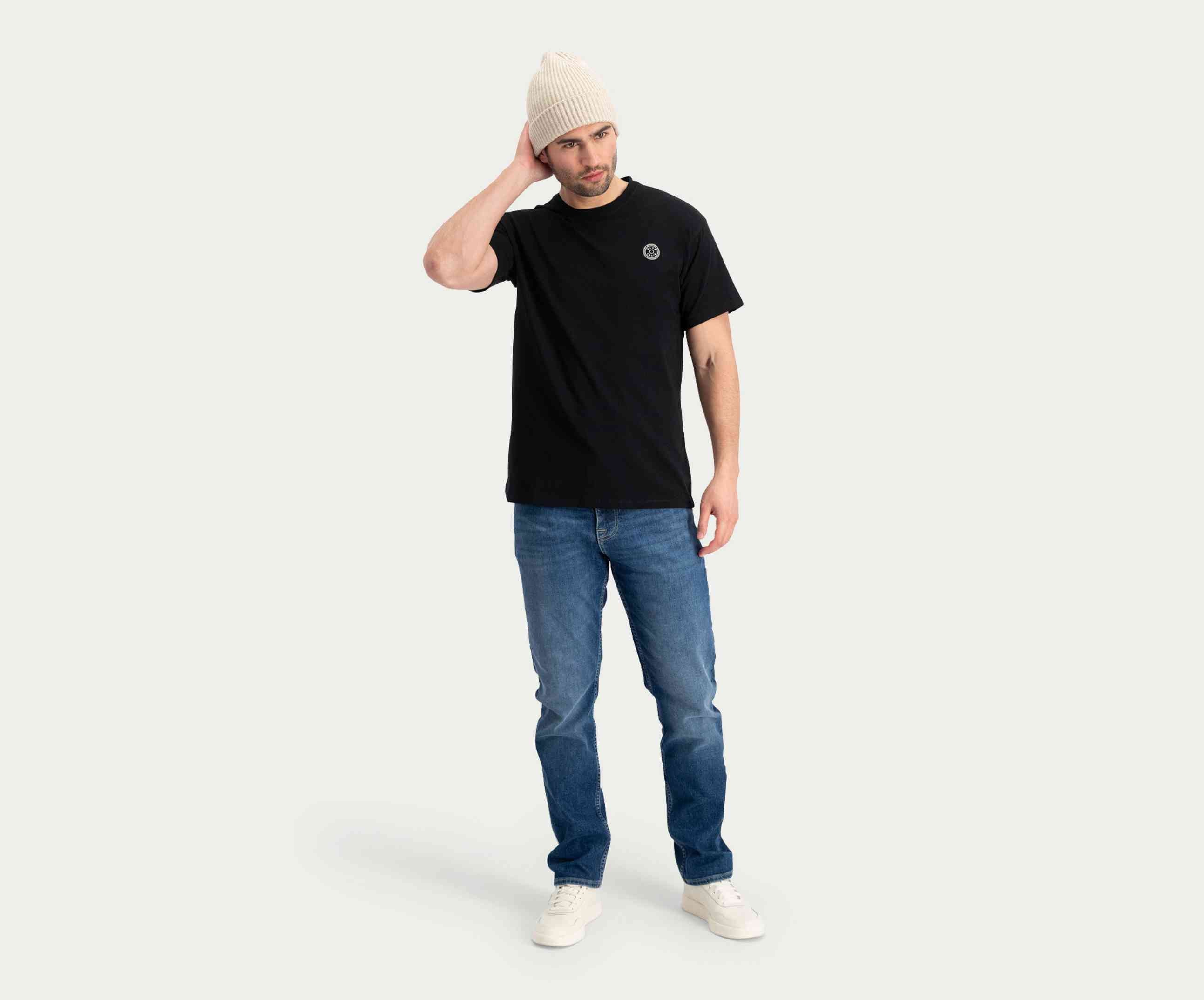 Een man draagt een zwart T-shirt, "The N01 Straight Indigo Used" van Blue Rivet Jeans, een witte muts en bijpassende schoenen en staat tegen een effen, lichte achtergrond. Hij houdt één hand op zijn hoofd en de andere rust naast hem.