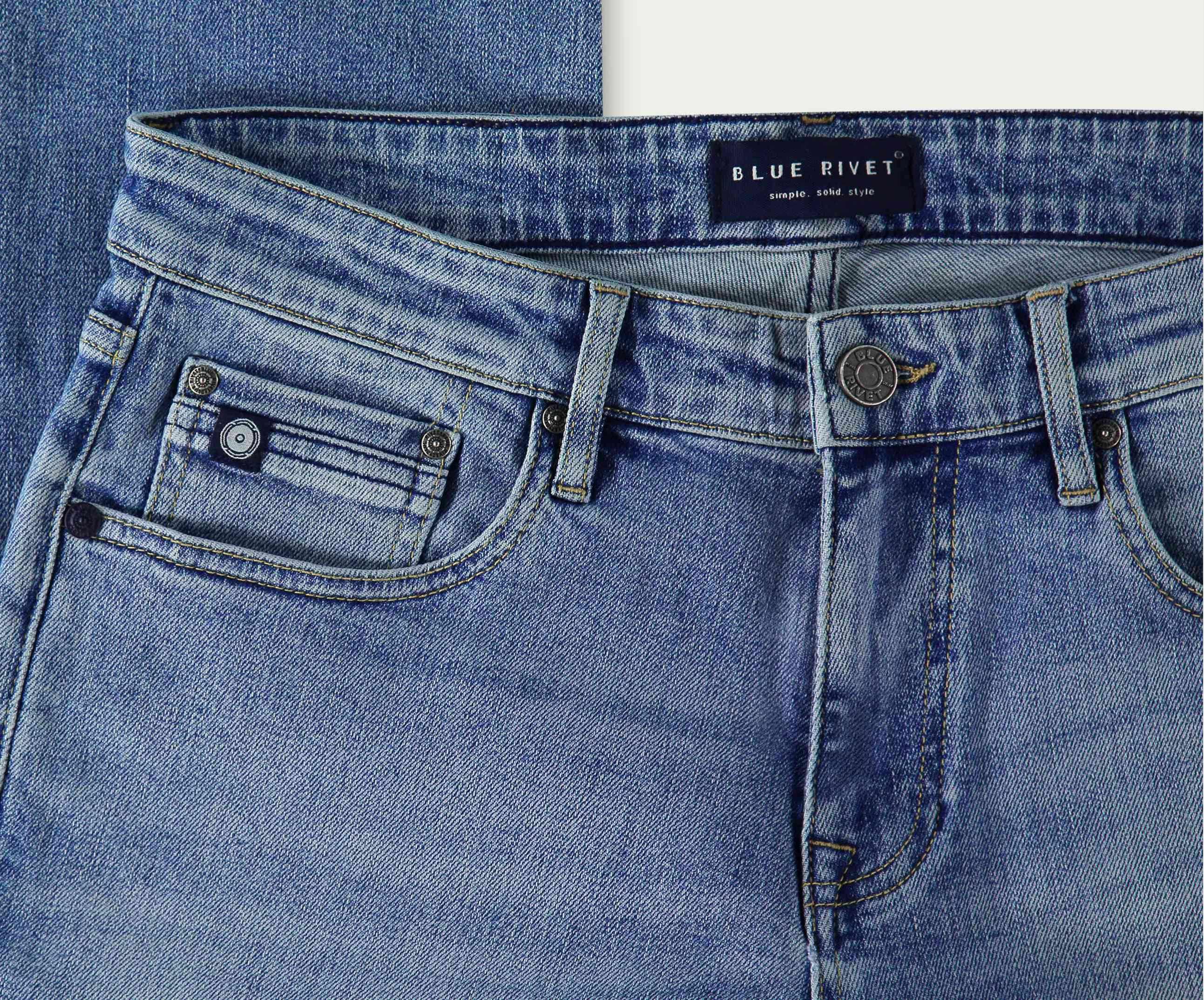 Close-up van lichtblauwe denim jeans met een Blue Rivet Jeans label. De N01 Straight Stone Bleached jeans heeft een knoopsluiting, zakken aan de voorkant en een klein muntzakje met een metalen klinknageldetail. Perfect ontworpen voor comfort en stijl, de stretchstof zorgt voor bewegingsgemak de hele dag door.