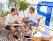 Twee mannen zitten op rieten stoelen op een balkon, in gesprek. Ze zijn gekleed in casual zomerkleding, waaronder The N01 Short Blue Black van Blue Rivet Jeans. Achter hen is een groot glazen raam met een blauwe letter P erop. In de buurt staat een potplant en een ronde houten tafel met kleine kopjes erop.