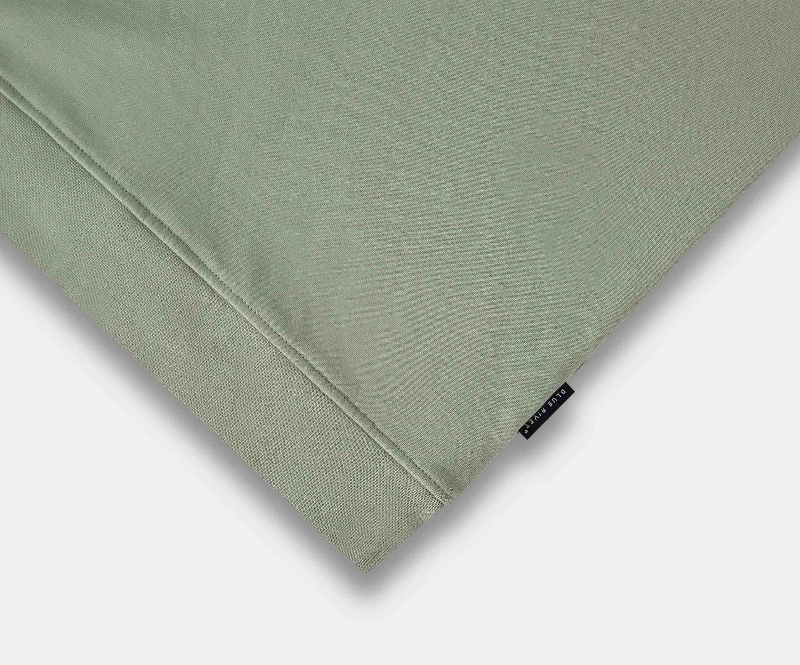 De afbeelding toont een close-up van een gevouwen stof van The N01 Sweat Seagrass van Blue Rivet Jeans. Het is gemaakt van biologisch katoen in een lichtgroene kleur, met een klein zwart label met witte tekst aan de rechterrand. Het gladde maar licht gestructureerde materiaal belichaamt de kwaliteit van deze premium trui hier.