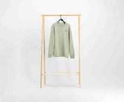 Een lichtgroene trui van Blue Rivet Jeans, bekend als The N01 Sweat Seagrass, gemaakt van biologisch katoen, hangt aan een zwarte hanger. Hij hangt op een houten rek tegen een effen witte achtergrond en heeft een normale pasvorm met een klein cirkelvormig ontwerp aan de linkerbovenkant.