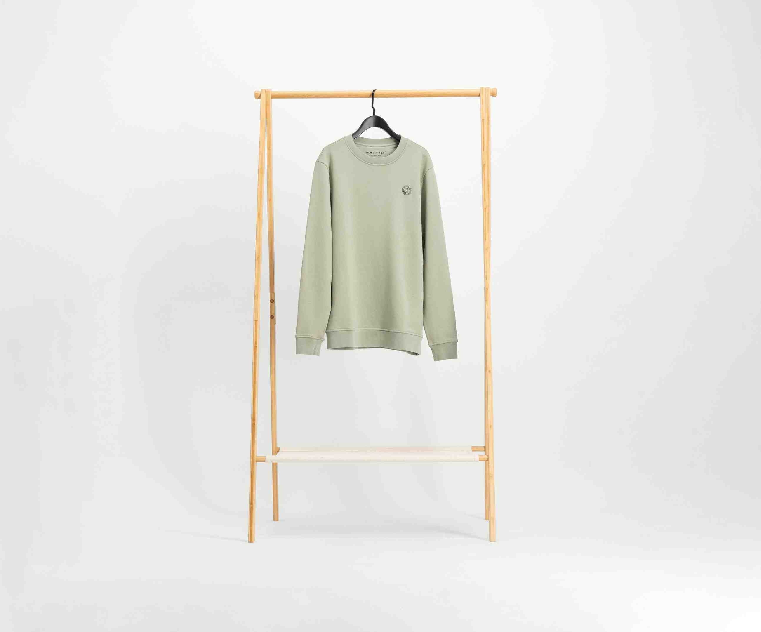 Een lichtgroene trui van Blue Rivet Jeans, bekend als The N01 Sweat Seagrass, gemaakt van biologisch katoen, hangt aan een zwarte hanger. Hij hangt op een houten rek tegen een effen witte achtergrond en heeft een normale pasvorm met een klein cirkelvormig ontwerp aan de linkerbovenkant.