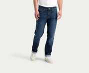 Op de afbeelding is een persoon te zien die "The N01 Straight Dark Indigo Used" van Blue Rivet Jeans draagt, gecombineerd met een wit T-shirt en witte sneakers. Het onderlichaam is zichtbaar tegen een lichtgrijze achtergrond.