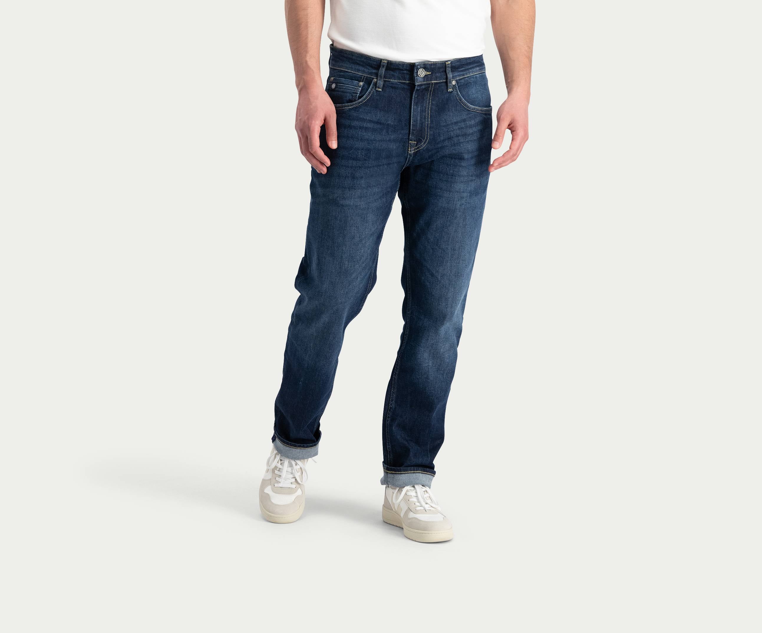 Op de afbeelding is een persoon te zien die "The N01 Straight Dark Indigo Used" van Blue Rivet Jeans draagt, gecombineerd met een wit T-shirt en witte sneakers. Het onderlichaam is zichtbaar tegen een lichtgrijze achtergrond.
