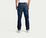 Een persoon staat met zijn rug naar de camera en draagt de N01 Straight Dark Indigo Used jeans van Blue Rivet Jeans, gestyled met opgerolde manchetten en een bruine leren patch op de tailleband. Ze worden gecombineerd met een strak wit shirt en witte sneakers tegen een effen lichtgrijze achtergrond.