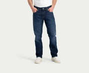 Een persoon draagt de N01 Straight Dark Indigo Used jeans van Blue Rivet Jeans, een wit overhemd en witte sneakers en staat tegen een effen achtergrond met zijn handen in zijn zakken.