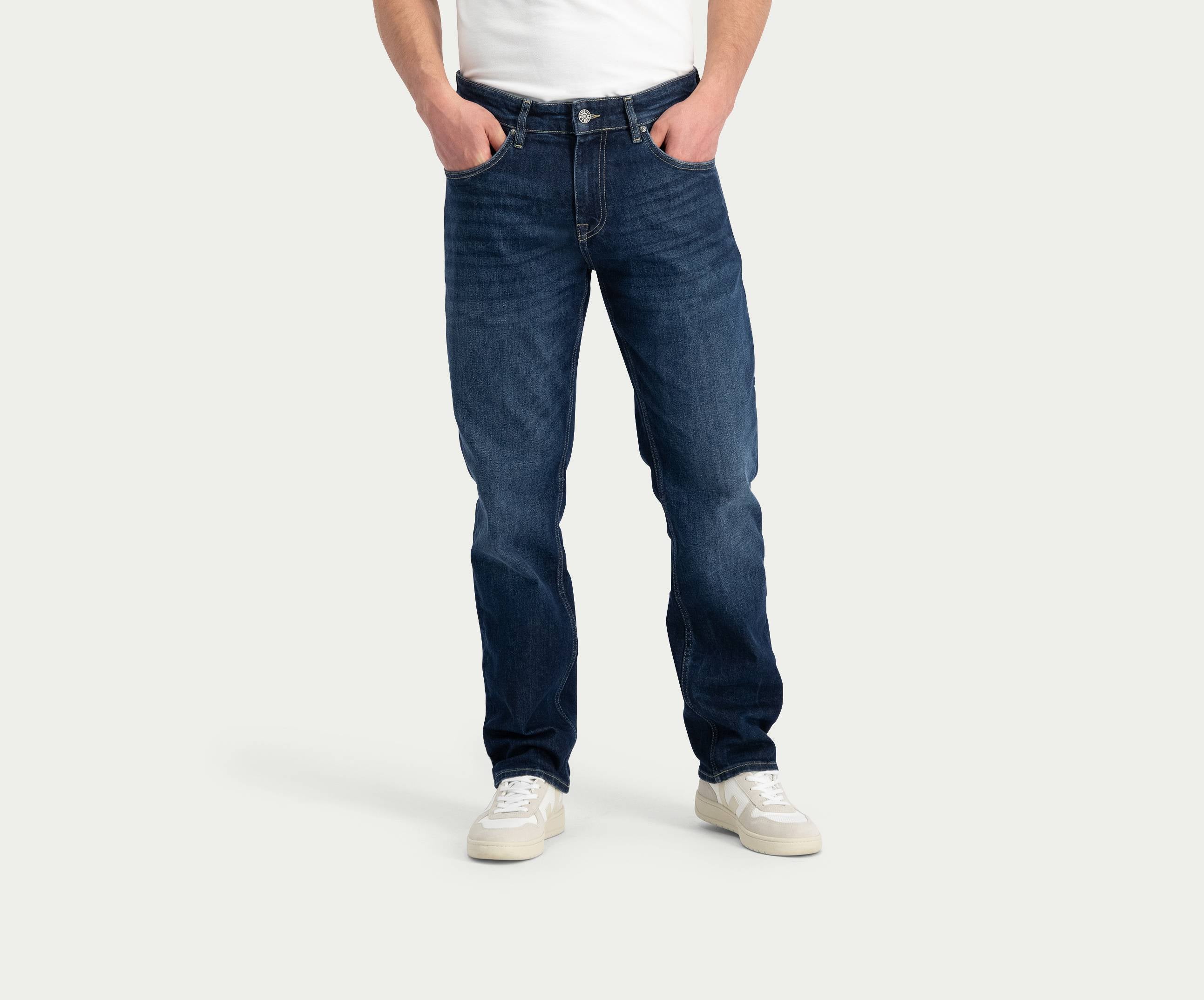 Een persoon draagt de N01 Straight Dark Indigo Used jeans van Blue Rivet Jeans, een wit overhemd en witte sneakers en staat tegen een effen achtergrond met zijn handen in zijn zakken.