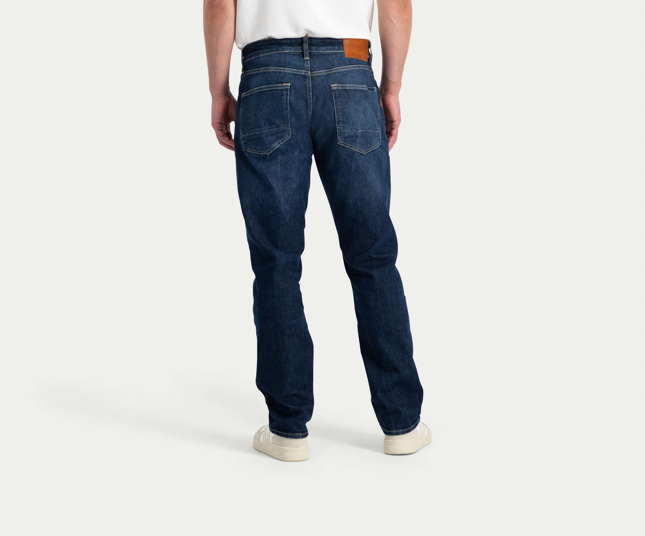 Een persoon is gekleed in Blue Rivet Jeans' "The N01 Straight Dark Indigo Used," gecombineerd met een wit T-shirt en witte sneakers. De jeans, van achteren gezien, benadrukken twee achterzakken en een bruine patch op de tailleband.