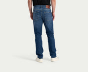 Een persoon die een Blue Rivet Jeans' The N01 Straight Indigo Used broek en een zwart shirt draagt, staat met zijn gezicht van de camera af en maakt zijn outfit compleet met witte sneakers. De lichtgekleurde, effen achtergrond accentueert de casual maar stijlvolle kleding.
