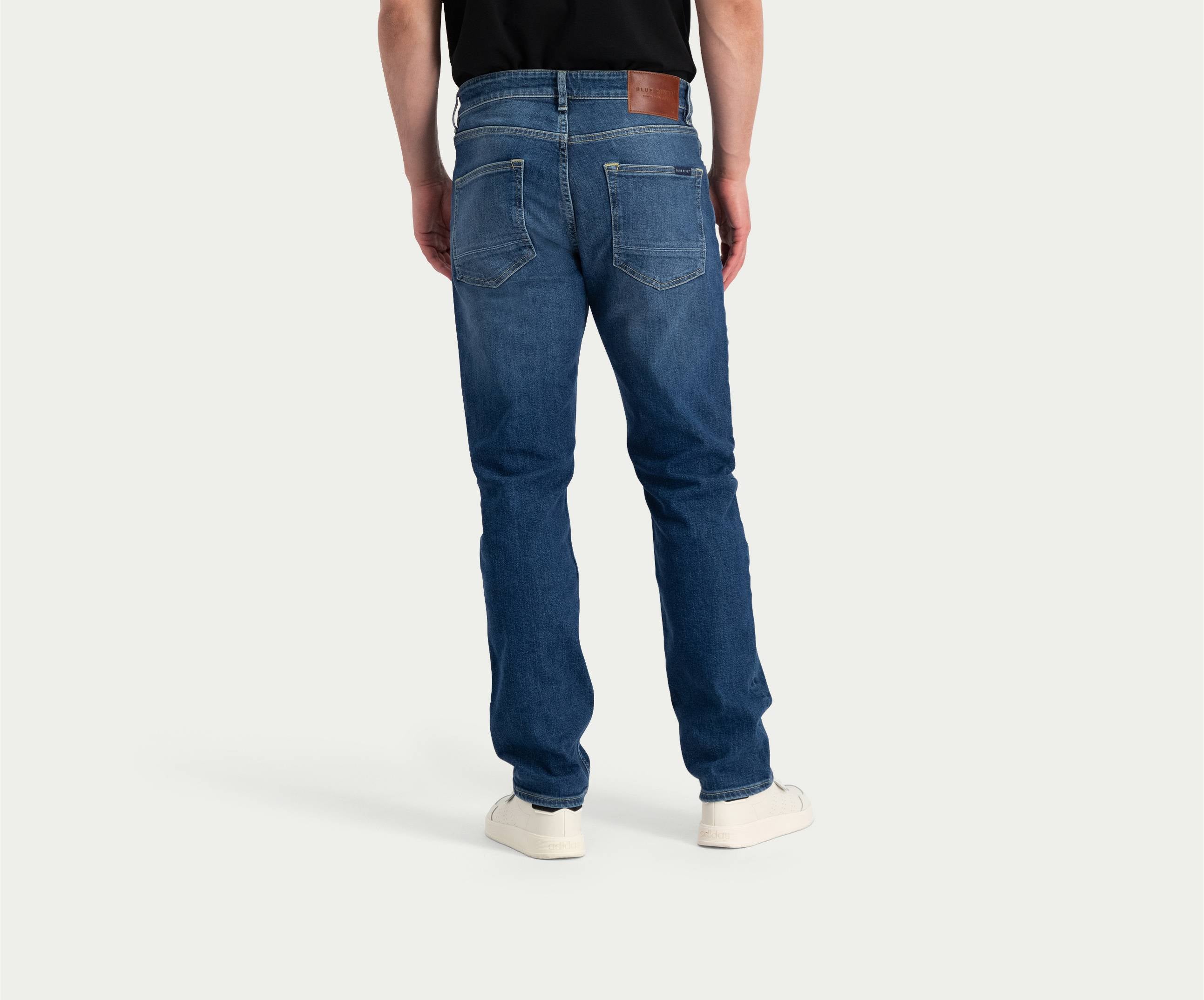 Een persoon die een Blue Rivet Jeans' The N01 Straight Indigo Used broek en een zwart shirt draagt, staat met zijn gezicht van de camera af en maakt zijn outfit compleet met witte sneakers. De lichtgekleurde, effen achtergrond accentueert de casual maar stijlvolle kleding.
