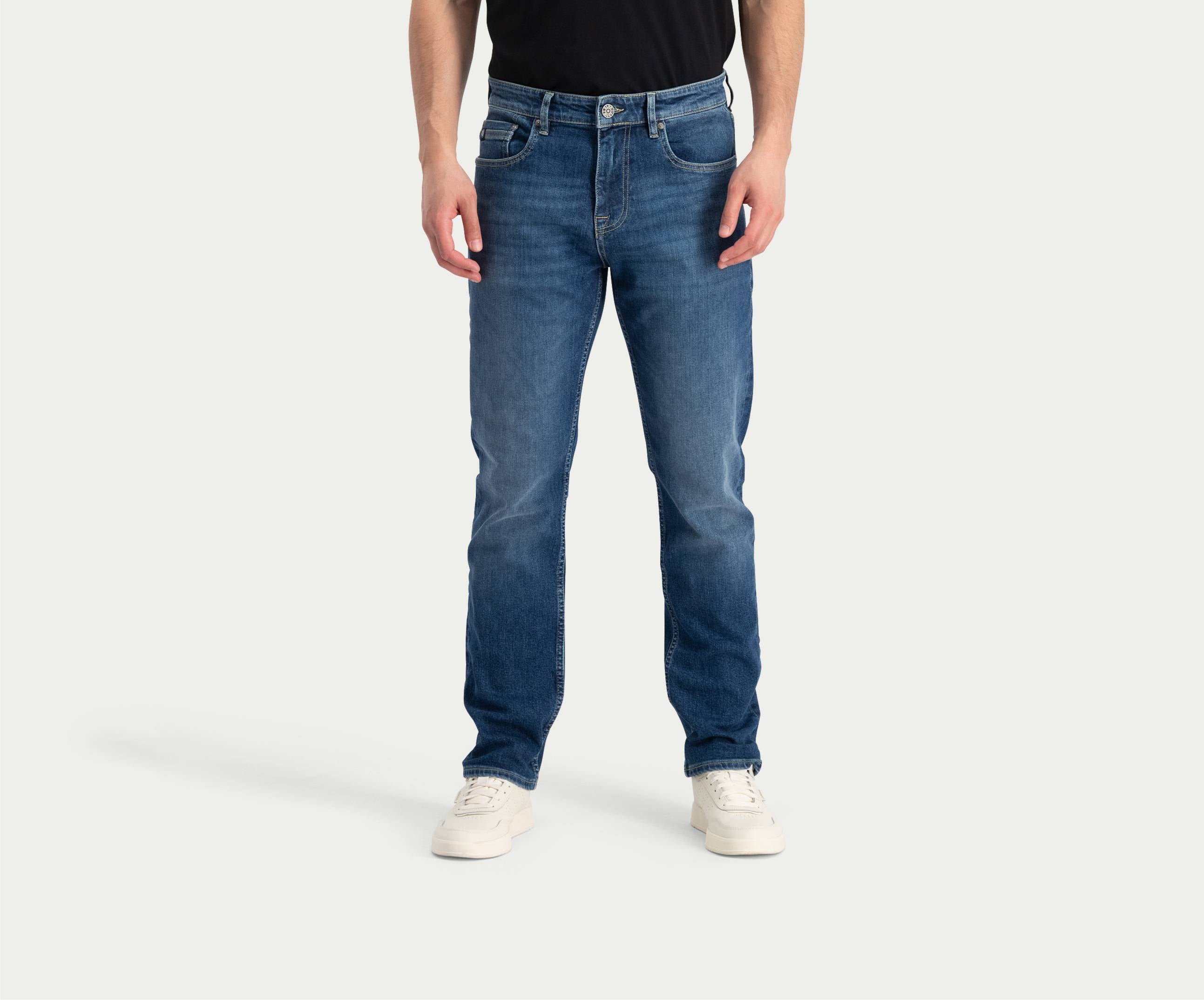 Een persoon die een zwart shirt draagt, de Blue Rivet Jeans' "The N01 Straight Indigo Used" met lichte vervaging, en witte sneakers staat tegen een effen achtergrond. Het hoofd van de persoon is niet zichtbaar op de afbeelding.