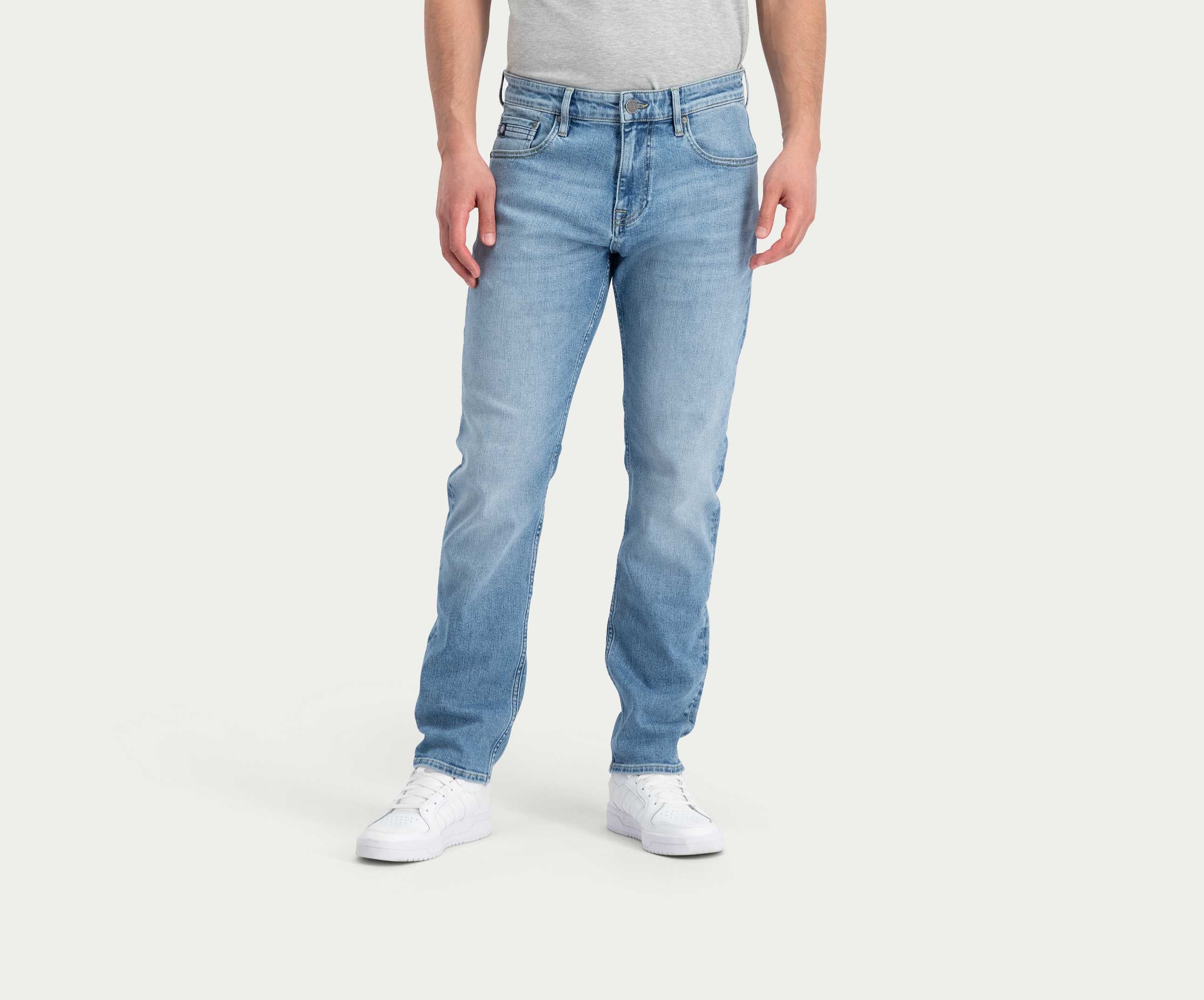 Een persoon die de N01 Straight Stone Bleached jeans van Blue Rivet Jeans en witte sneakers draagt, staat tegen een effen achtergrond. De stretchjeans heeft een klassieke snit en de persoon draagt ook een lichtgrijs t-shirt. De afbeelding richt zich op het onderlichaam.