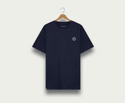 Een marineblauwe The N01 Tee van Blue Rivet Jeans hangt aan een houten hanger tegen een lichte achtergrond. Dit regular fit shirt heeft een klein rond embleem op de linkerborst en een contrasterend halsdetail.