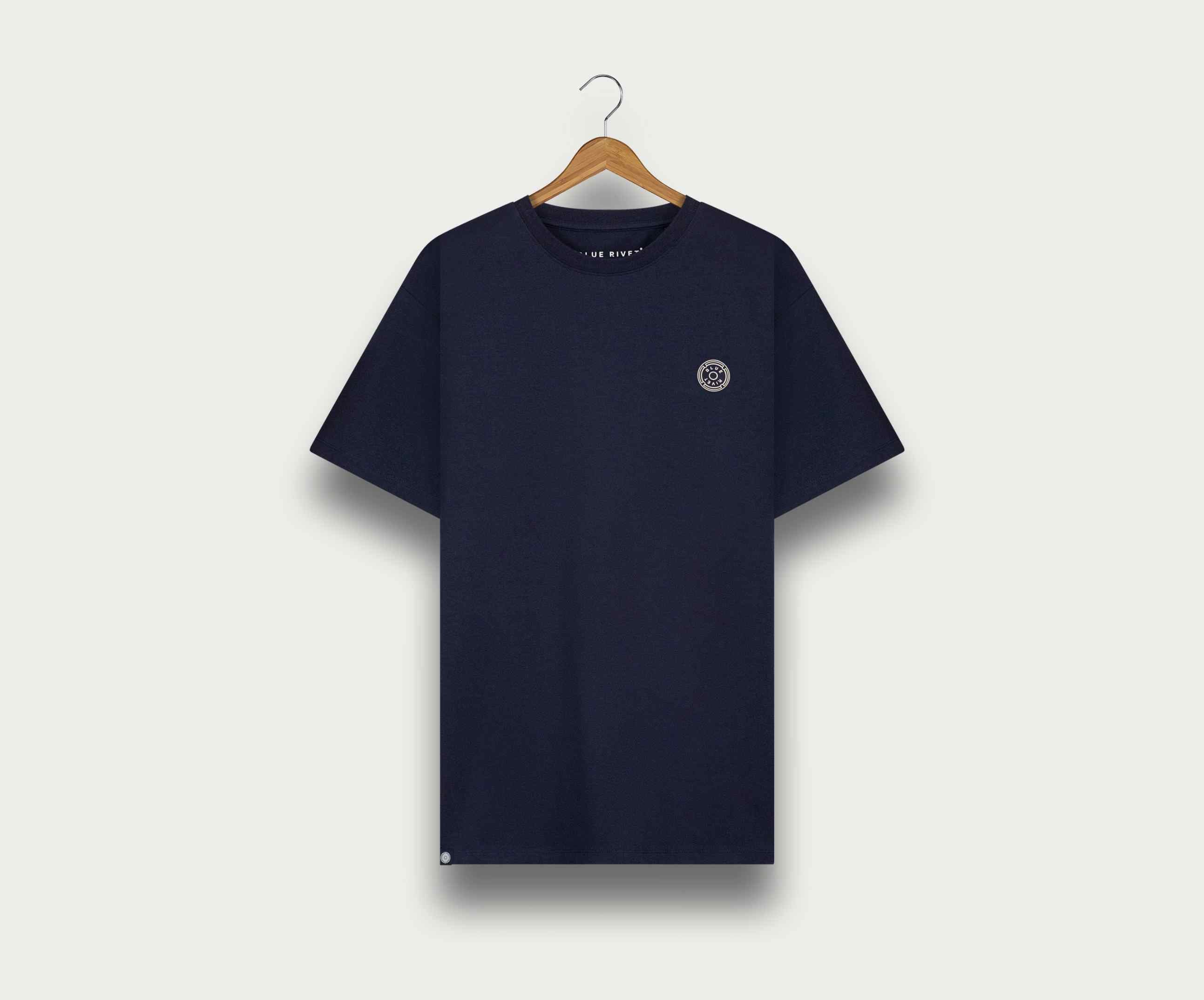 Een marineblauwe The N01 Tee van Blue Rivet Jeans hangt aan een houten hanger tegen een lichte achtergrond. Dit regular fit shirt heeft een klein rond embleem op de linkerborst en een contrasterend halsdetail.