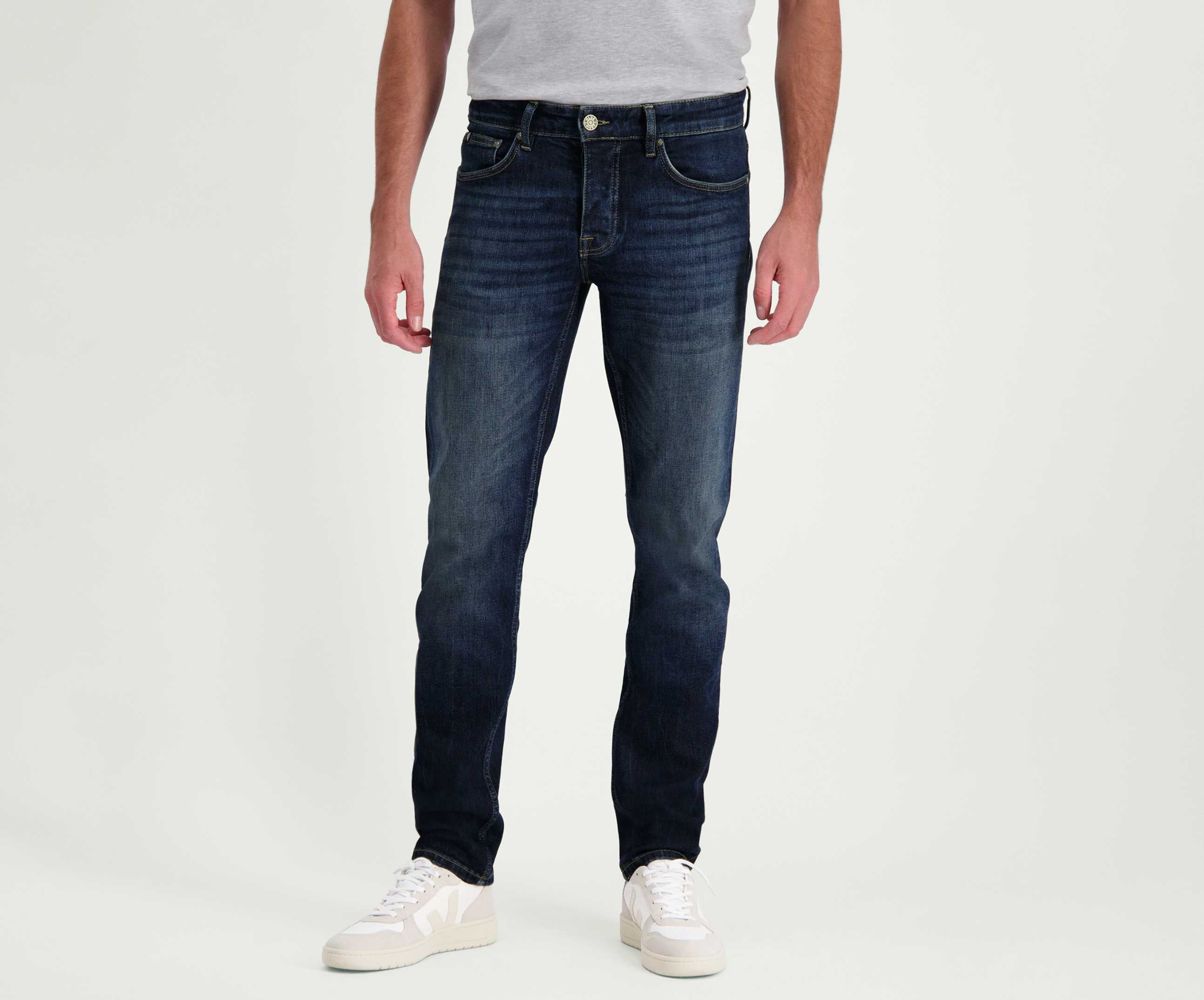 Voor een effen witte achtergrond draagt een individu een grijs t-shirt gecombineerd met Blue Rivet Jeans' The N01 Slim Dark Indigo Used denim, met een licht vervaagde textuur. Het ensemble wordt gecompleteerd met witte sneakers, met de focus op de premium denim stijl omdat het bovenlichaam uit het zicht blijft.