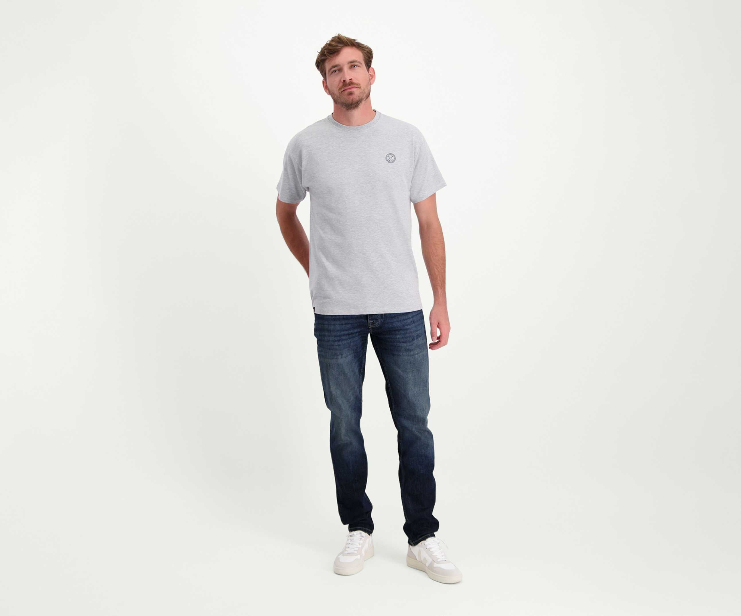 Een man draagt een grijs T-shirt en The N01 Slim Dark Indigo Used by Blue Rivet Jeans en staat tegen een effen witte achtergrond, met één hand subtiel op zijn rug gestrekt.