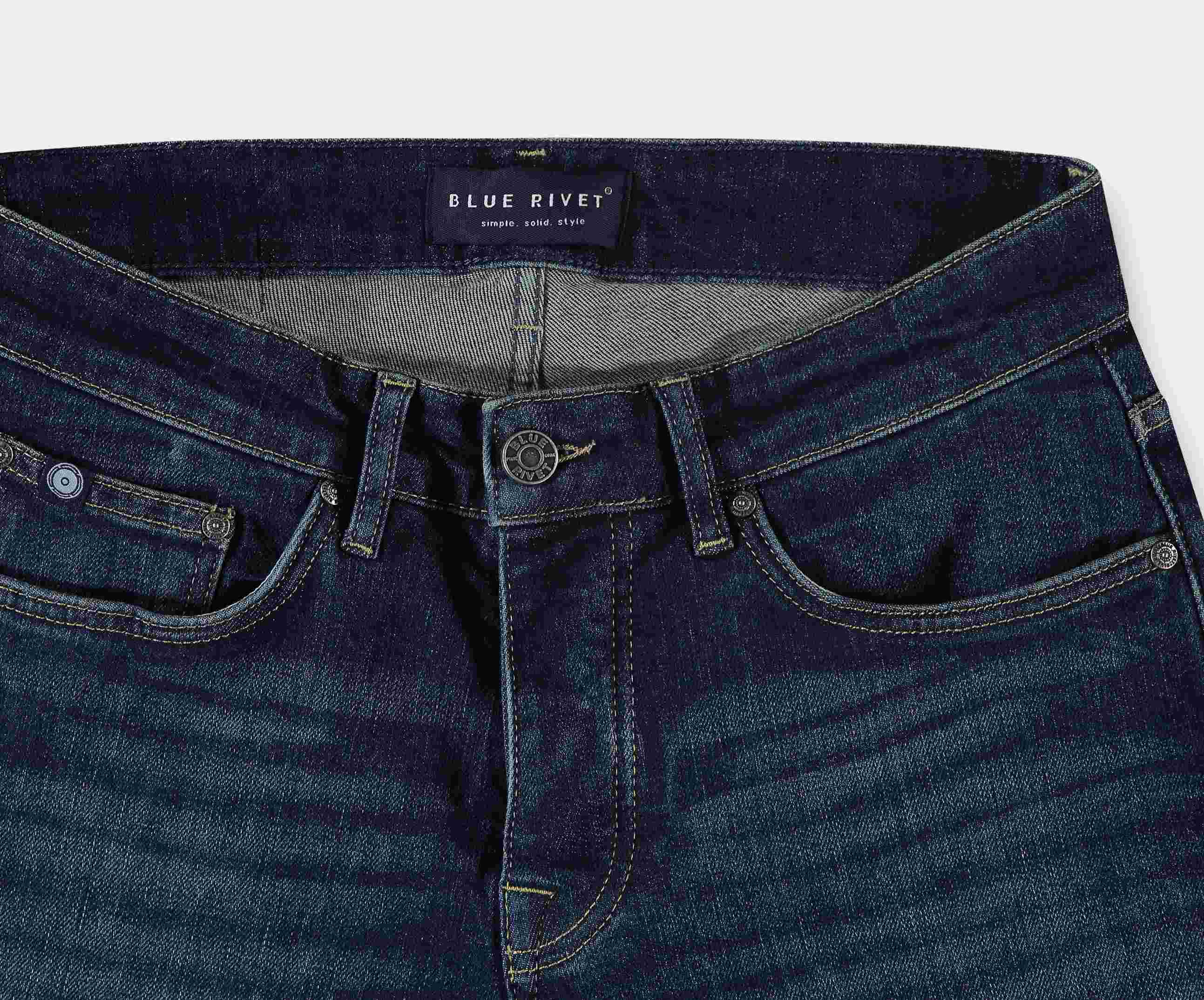 Close-up van een paar The N01 Slim Dark Indigo Used denim jeans met een zwart label op de tailleband met de tekst Blue Rivet Jeans. Deze stretchjeans hebben een gulp met knoop en rits, zakken aan de voorkant en een licht vervaagde textuur.