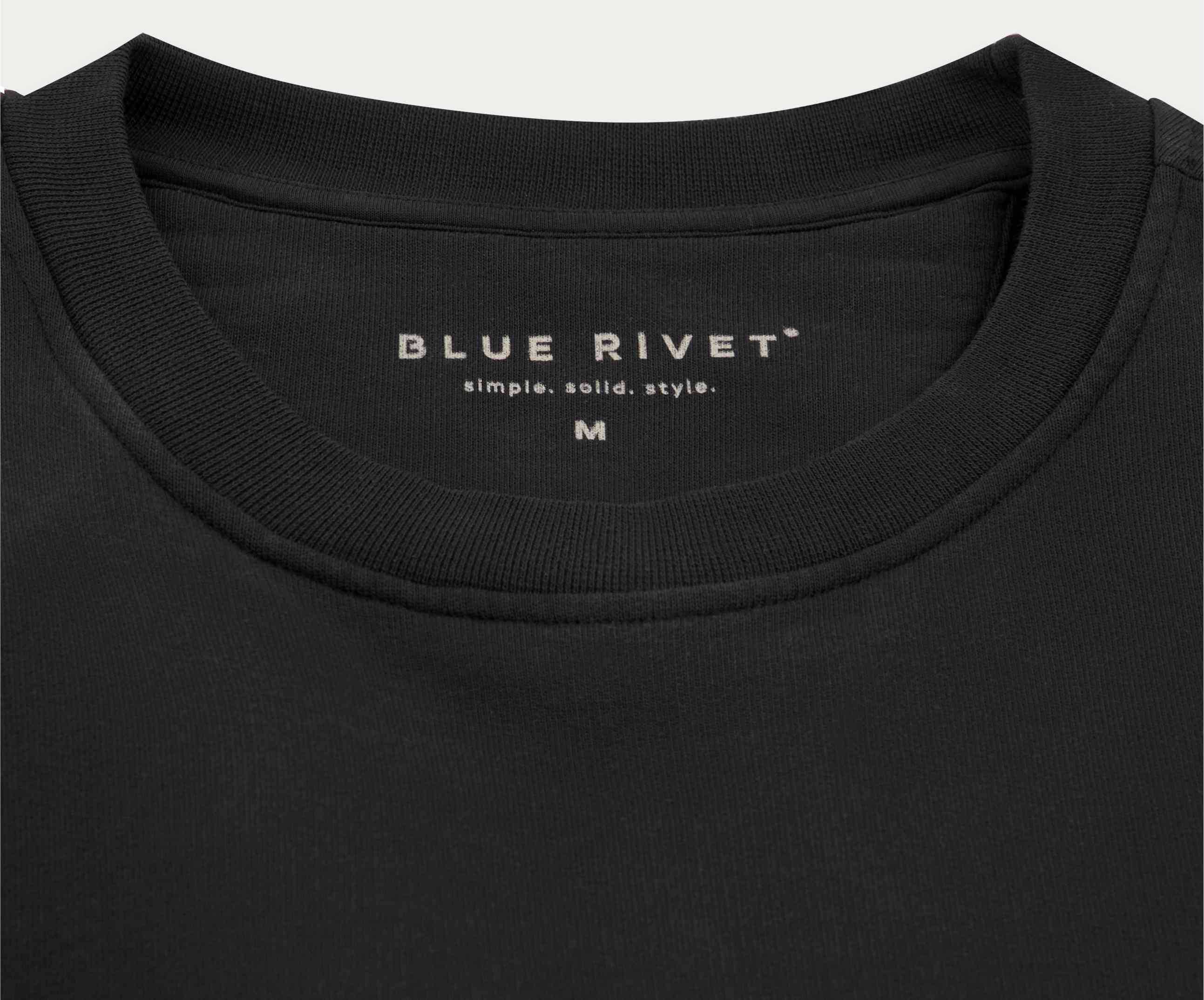 Close-up van een zwart T-shirt met het label Blue Rivet Jeans boven hun slogan simple. solid. style., met maat M op de binnenkant van de kraag. Gemaakt van biologisch katoen, dit zachte en nauwsluitende stuk is gemaakt voor degenen die premium kwaliteit waarderen, verwant aan onze geliefde The N01 Sweat Anthracite-lijn.