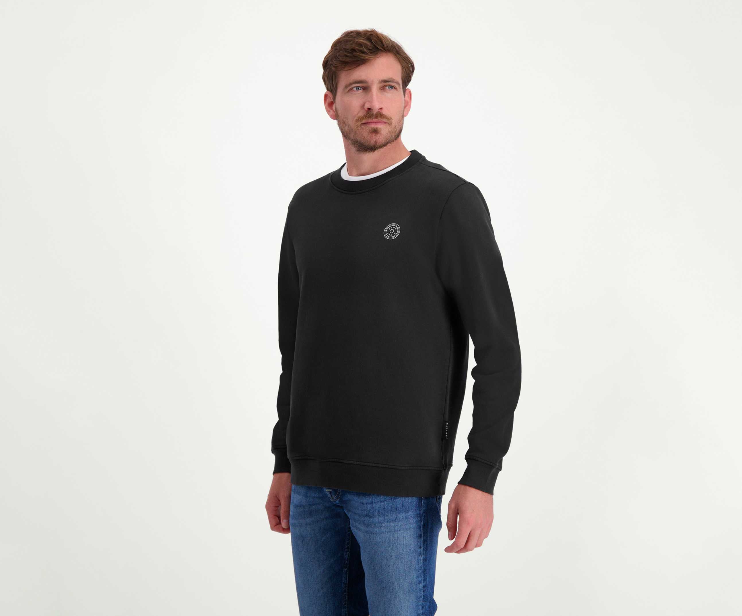 Een man met kort bruin haar en een baard draagt The N01 Sweat Anthracite van het merk Blue Rivet Jeans, gecombineerd met een regular fit blauwe jeans. Hij staat tegen een effen witte achtergrond en kijkt lichtjes naar links. De sweater heeft een klein rond logo op de borst.