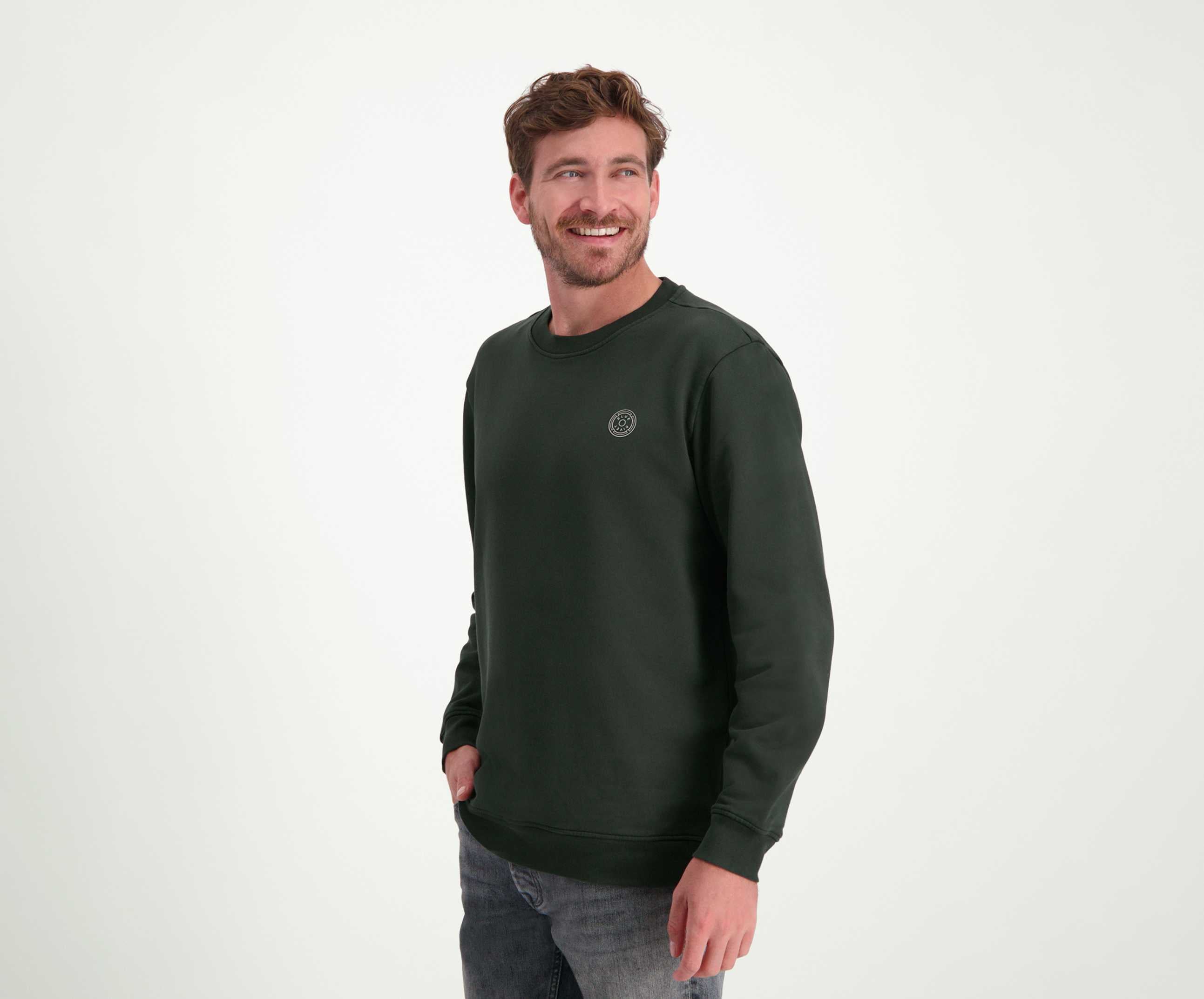 Een lachende man met kort bruin haar draagt The N01 Sweat Dark Green van Blue Rivet Jeans, gemaakt van biologisch katoen, en grijze jeans. Hij staat tegen een effen witte achtergrond met zijn handen in zijn zakken, en kijkt lichtjes opzij.