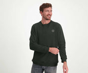 Een persoon met kort bruin haar en een baard glimlacht terwijl hij opzij kijkt. Ze dragen The N01 Sweat Dark Green van Blue Rivet Jeans, gecombineerd met jeans, tegen een effen witte achtergrond.