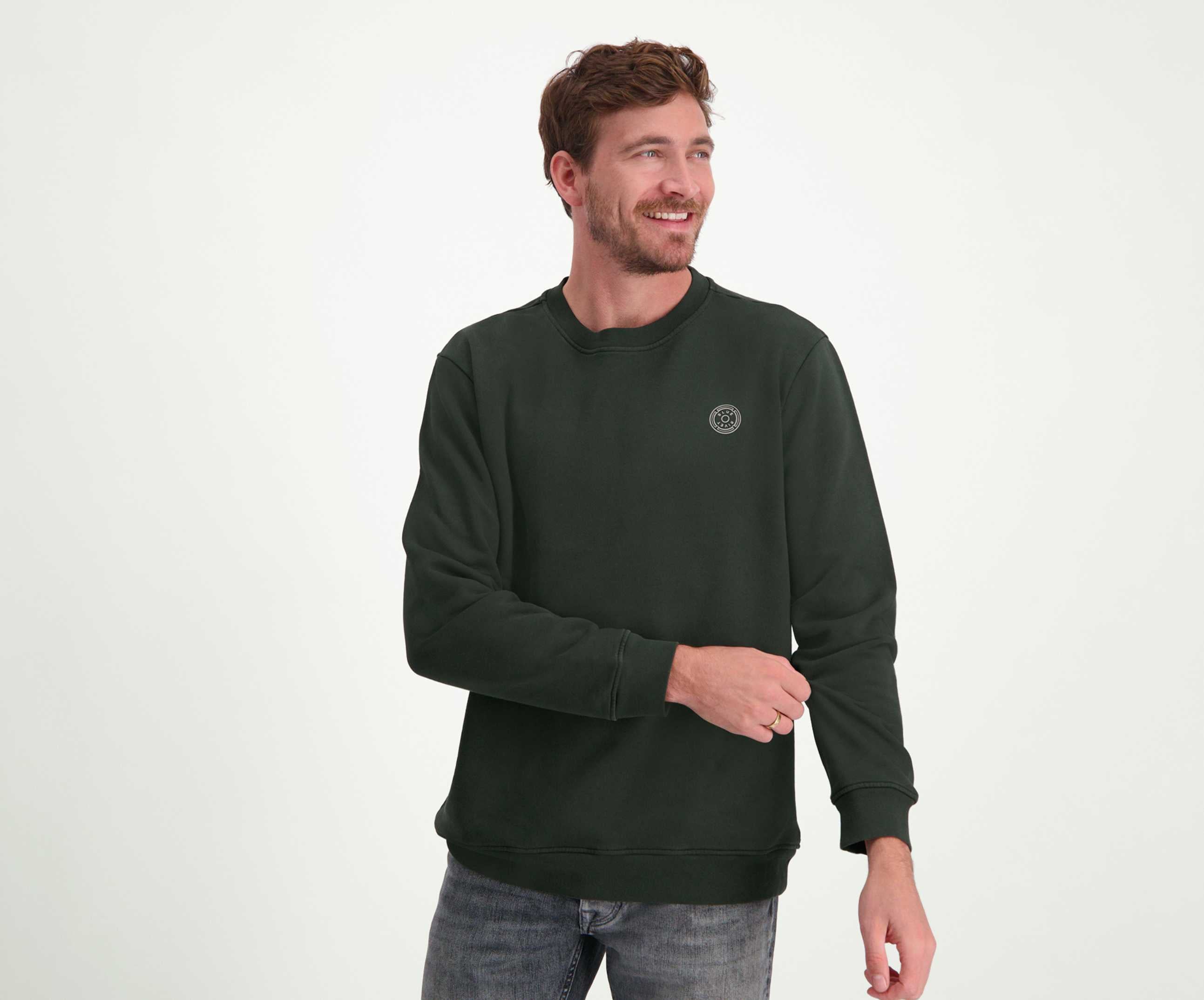 Een persoon met kort bruin haar en een baard glimlacht terwijl hij opzij kijkt. Ze dragen The N01 Sweat Dark Green van Blue Rivet Jeans, gecombineerd met jeans, tegen een effen witte achtergrond.