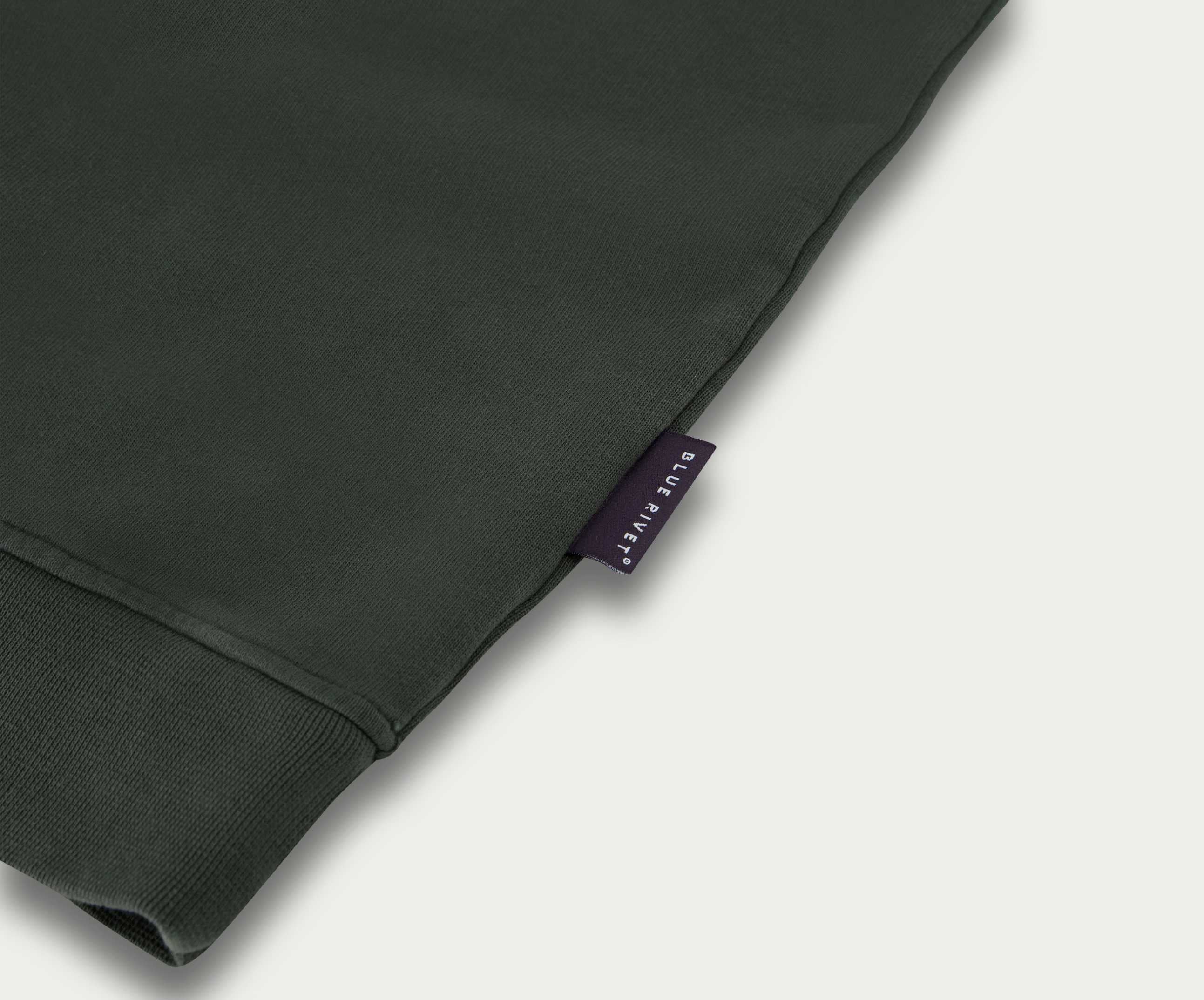 Close-up van een donkergroene stof van "The N01 Sweat Dark Green" van Blue Rivet Jeans, met een klein zwart label gestikt op de rand. De stof is gemaakt van biologisch katoen en heeft een manchetdetail, wat het verfijnde ontwerp en de milieuvriendelijke oorsprong weerspiegelt.