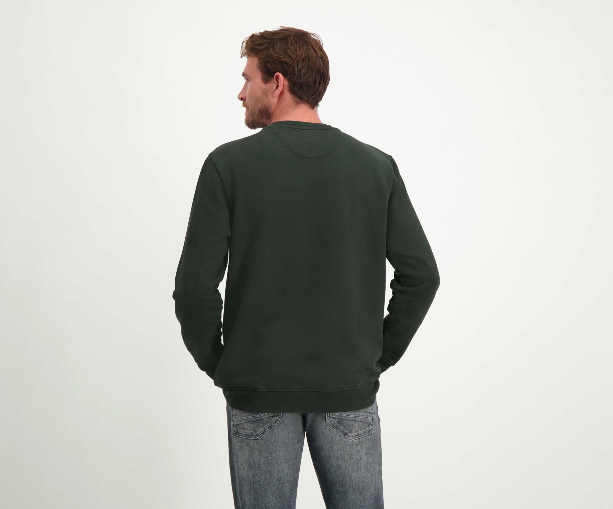 Een man met kort bruin haar staat met zijn rug naar de camera en draagt The N01 Sweat Dark Green van Blue Rivet Jeans, gemaakt van biologisch katoen. Deze donkergroene sweater combineert naadloos met zijn grijze jeans tegen een effen witte achtergrond.