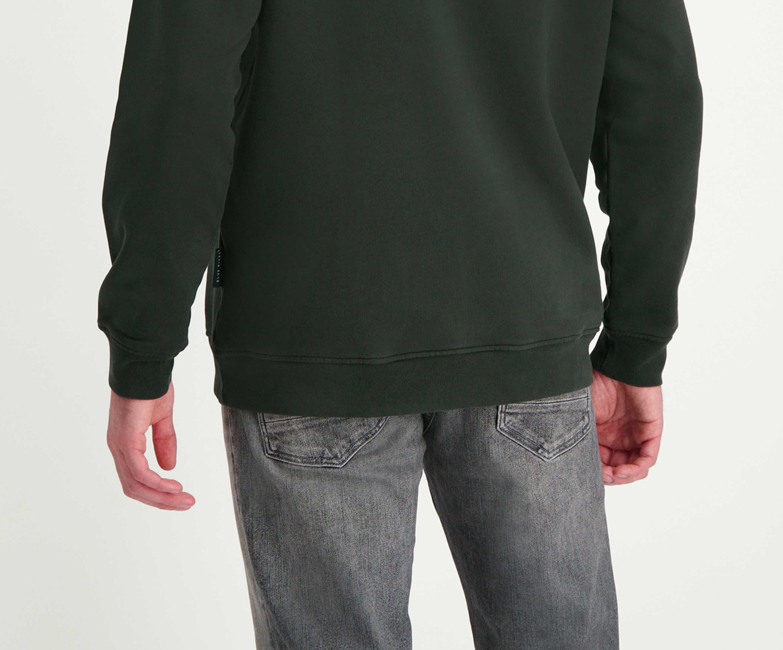 Een persoon is van achteren afgebeeld met de N01 Sweat Dark Green trui, een product van Blue Rivet Jeans, gemaakt van biologisch katoen. Ze dragen ook een grijze jeans in een normale pasvorm met zichtbare achterzakken. Hun linkerhand is gedeeltelijk zichtbaar, met vingers licht gekruld, tegen een effen witte achtergrond.