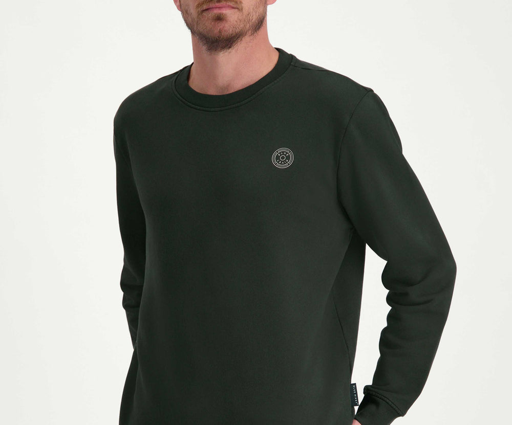 Een lachende man met kort bruin haar draagt The N01 Sweat Dark Green van Blue Rivet Jeans, gemaakt van biologisch katoen, en grijze jeans. Hij staat tegen een effen witte achtergrond met zijn handen in zijn zakken, en kijkt lichtjes opzij.