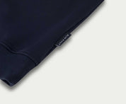 Close-up van een gevouwen marineblauwe The N01 Sweat Navy van Blue Rivet Jeans, gemaakt van biologisch katoen. De afbeelding benadrukt de mouw en een klein zwart label met witte tekst bij de rand, alles tegen een lichtgrijze achtergrond.