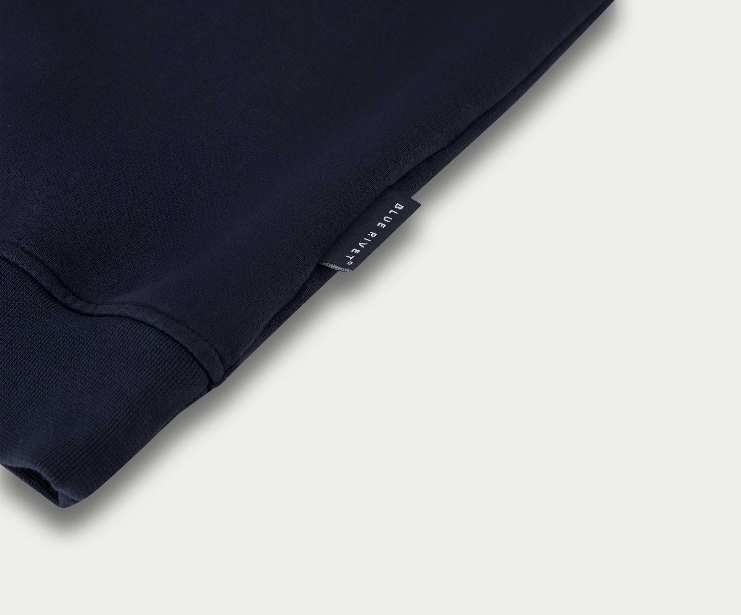 Close-up van een gevouwen marineblauwe The N01 Sweat Navy van Blue Rivet Jeans, gemaakt van biologisch katoen. De afbeelding benadrukt de mouw en een klein zwart label met witte tekst bij de rand, alles tegen een lichtgrijze achtergrond.