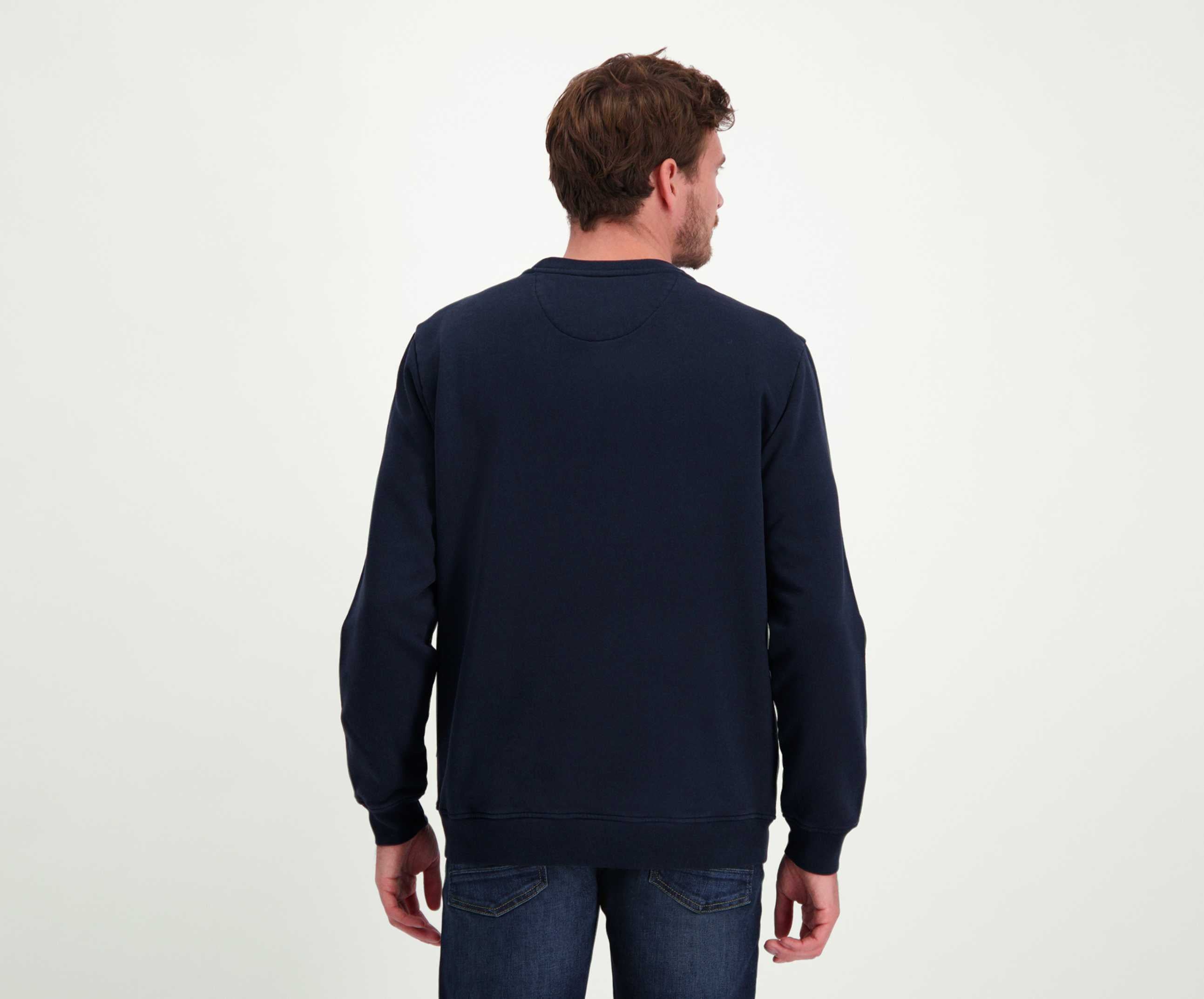 Een man met kort bruin haar staat en kijkt weg van de camera, en toont een regular fit The N01 Sweat Navy trui. Hij draagt Blue Rivet Jeans, en de setting is een effen lichtgrijze achtergrond.