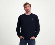 Een man met kort haar en een baard draagt The N01 Sweat Navy, een donkere, regular fit, long-sleeve trui van Blue Rivet Jeans. Hij is gemaakt van biologisch katoen en heeft een klein rond logo op de borst. Hij heeft hem gecombineerd met jeans en staat nonchalant met zijn handen in zijn zakken tegen een effen witte achtergrond.