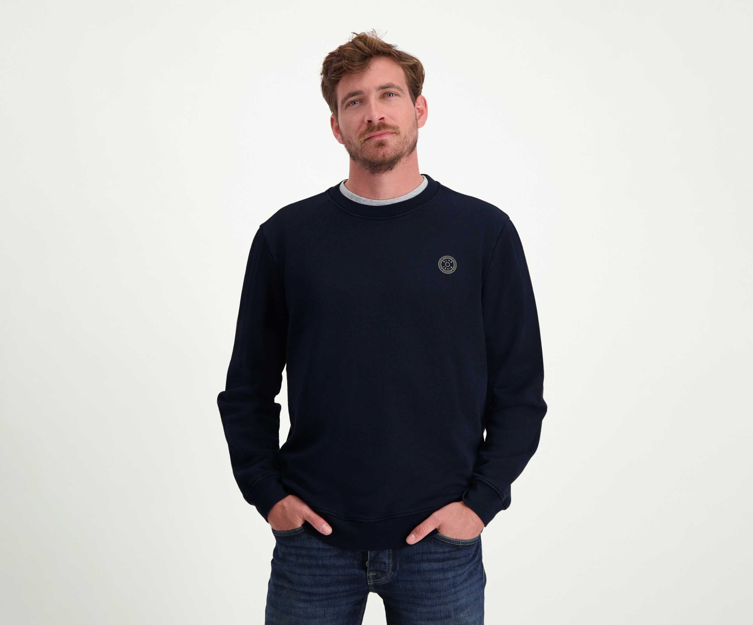 Een man met kort haar en een baard draagt The N01 Sweat Navy, een donkere, regular fit, long-sleeve trui van Blue Rivet Jeans. Hij is gemaakt van biologisch katoen en heeft een klein rond logo op de borst. Hij heeft hem gecombineerd met jeans en staat nonchalant met zijn handen in zijn zakken tegen een effen witte achtergrond.