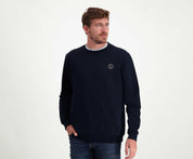 Een persoon met kort bruin haar en een baard draagt *The N01 Sweat Navy* van Blue Rivet Jeans, gemaakt van premium biologisch katoen in een normale pasvorm, gecombineerd met jeans. De persoon kijkt opzij met één hand in een zak, staande tegen een effen witte achtergrond.