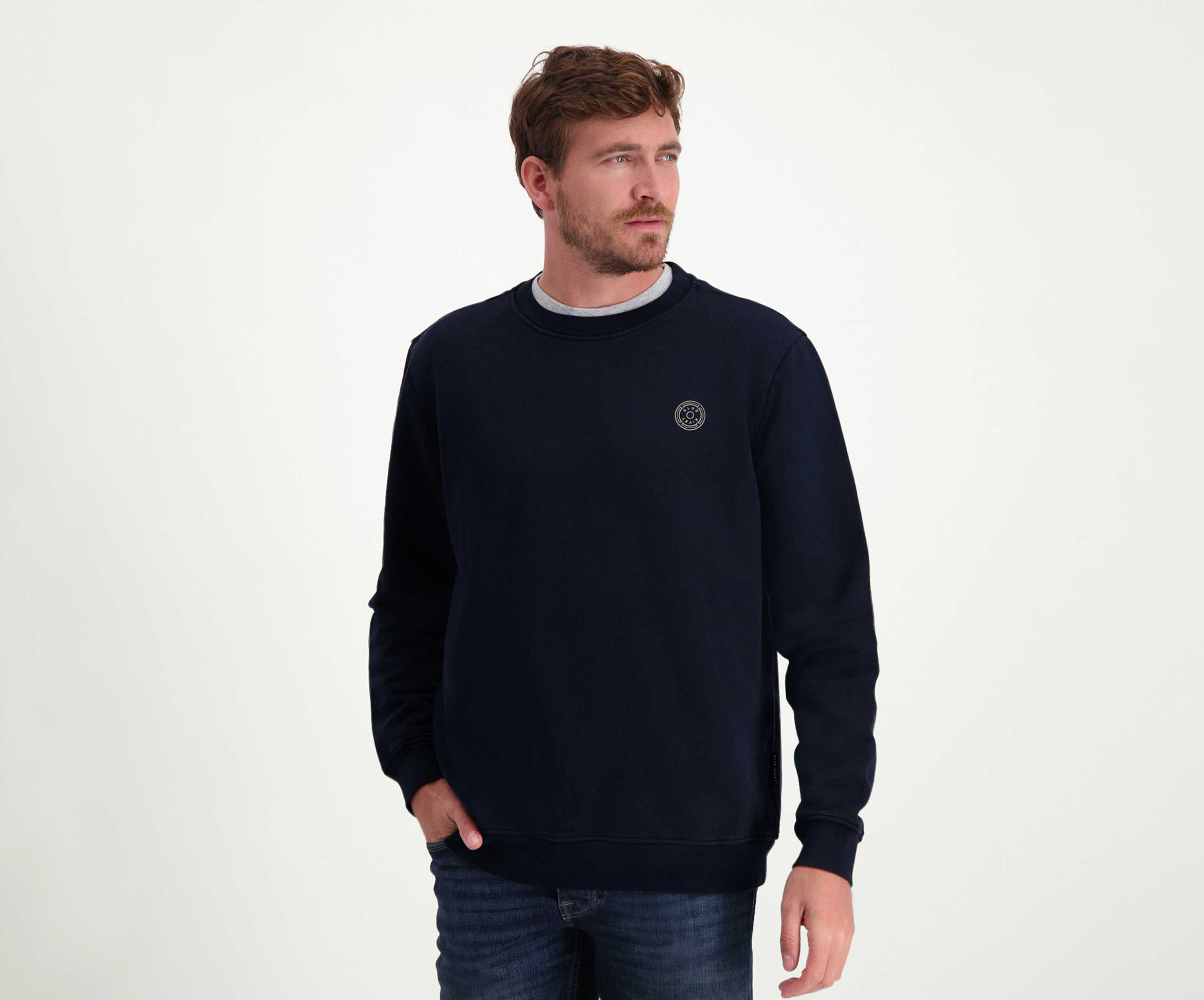 Een persoon met kort bruin haar en een baard draagt *The N01 Sweat Navy* van Blue Rivet Jeans, gemaakt van premium biologisch katoen in een normale pasvorm, gecombineerd met jeans. De persoon kijkt opzij met één hand in een zak, staande tegen een effen witte achtergrond.