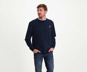 Een man met kort haar draagt The N01 Sweat Navy van Blue Rivet Jeans, gemaakt van biologisch katoen, naast een blauwe spijkerbroek. Hij staat met zijn handen in zijn zakken en kijkt zijwaarts tegen een effen, lichtgekleurde achtergrond die de normale pasvorm van de trui accentueert.